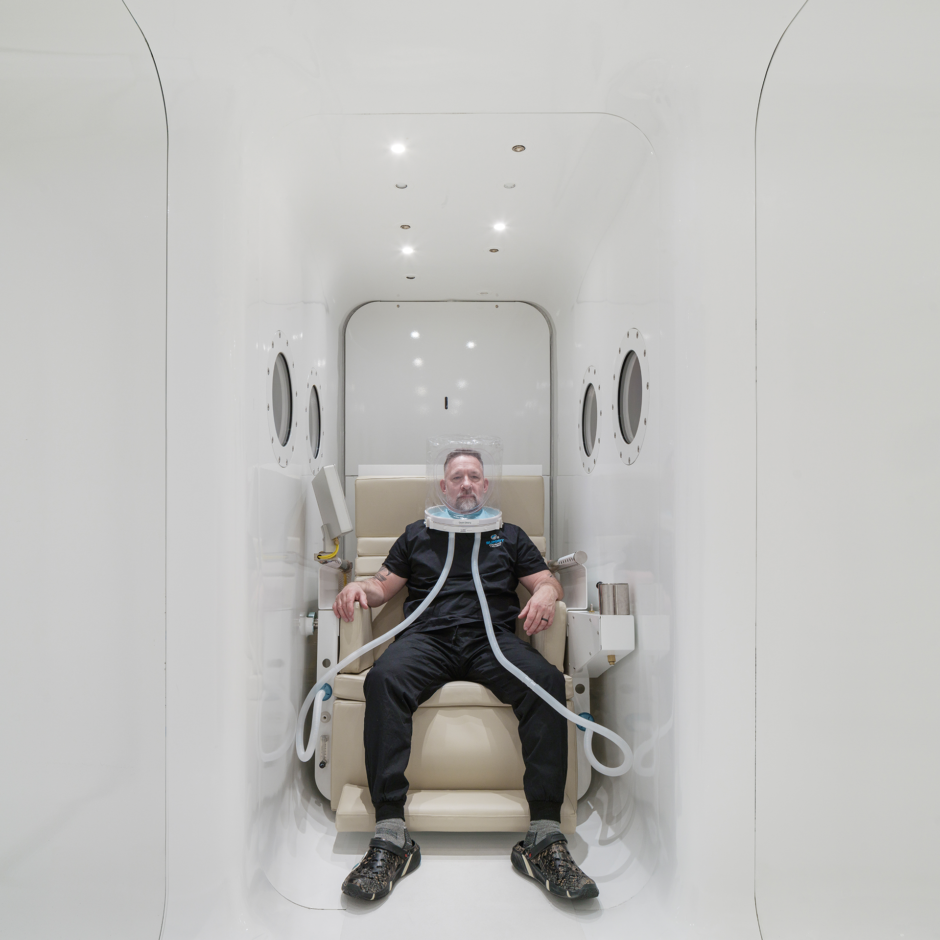 Director of Hyperbarics & Safety at Summit Hyperbarics & Wellness sitting in a chair inside the HBOT chamber, wearing an oxygen hood during a therapy session.