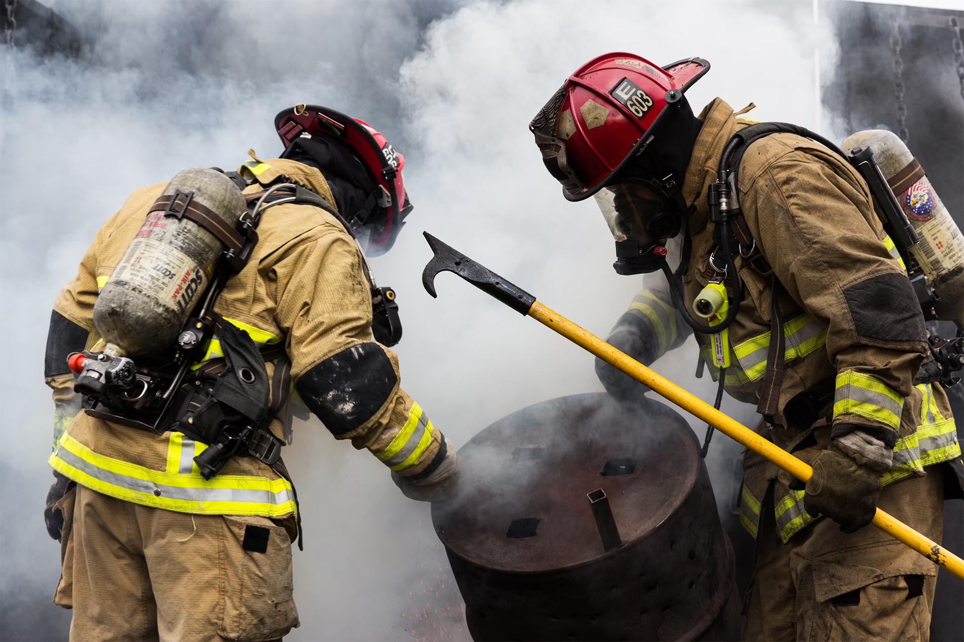 Two firefighters in full protective gear and oxygen tanks working through heavy smoke, using tools to manage a burning barrel.