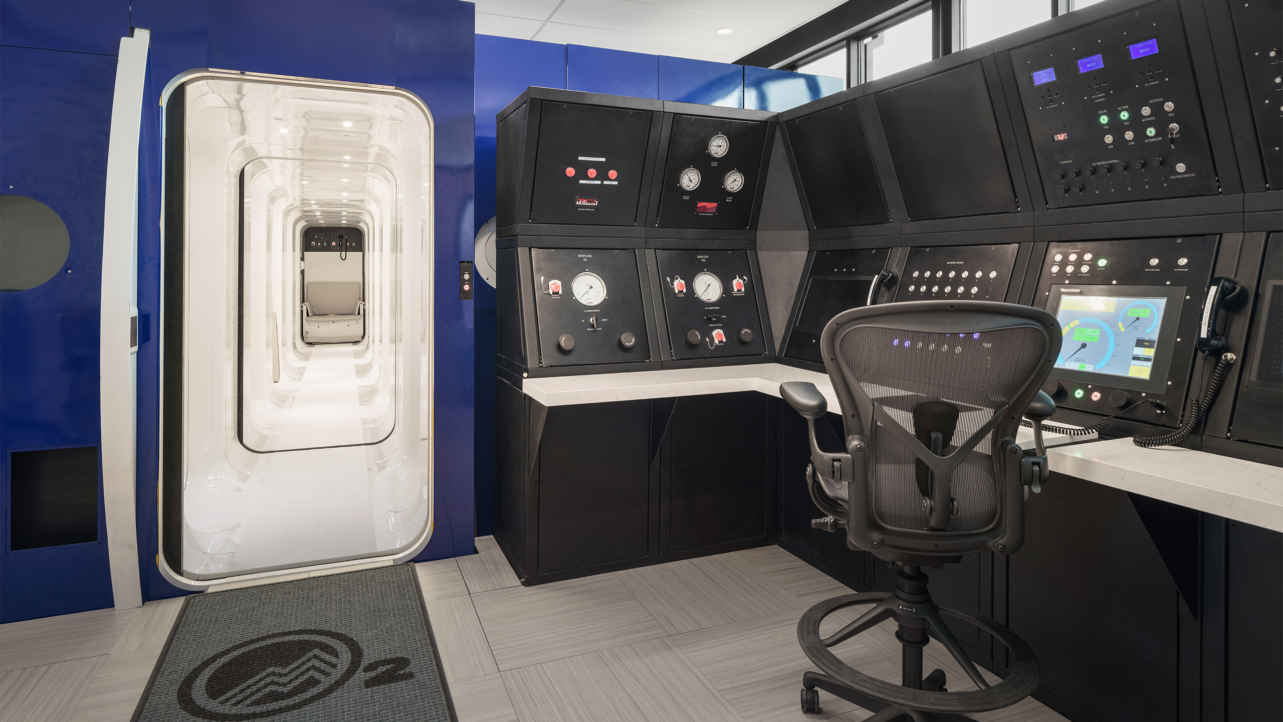 Entrance to a hyperbaric oxygen therapy chamber at Summit Hyperbarics & Wellness, featuring a sleek white interior and a control panel in the adjacent operator's station.