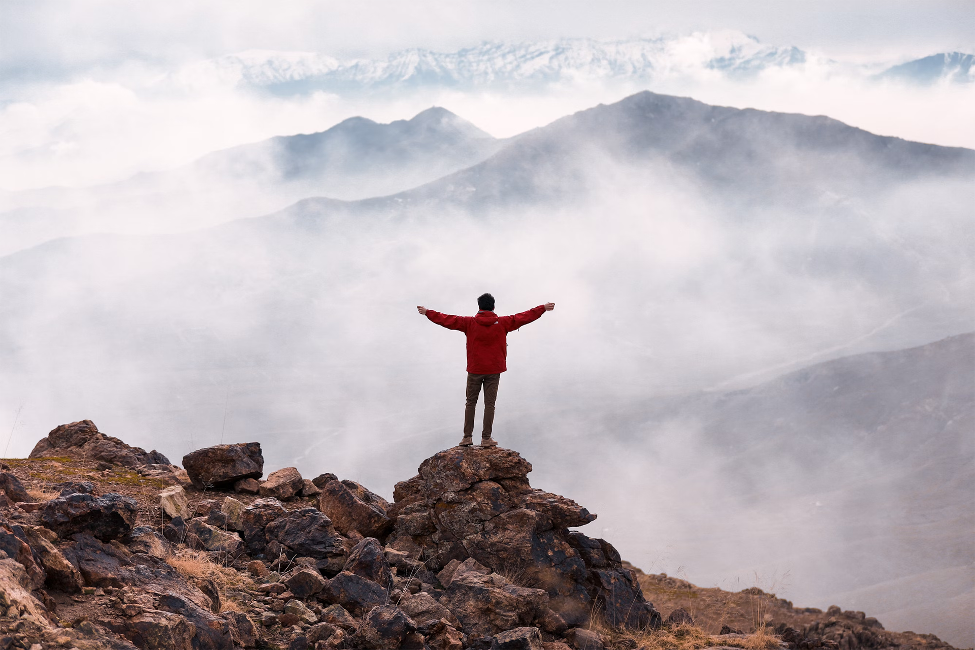 A person in a red jacket standing on a rocky peak with arms outstretched, overlooking misty mountain ranges in the background.